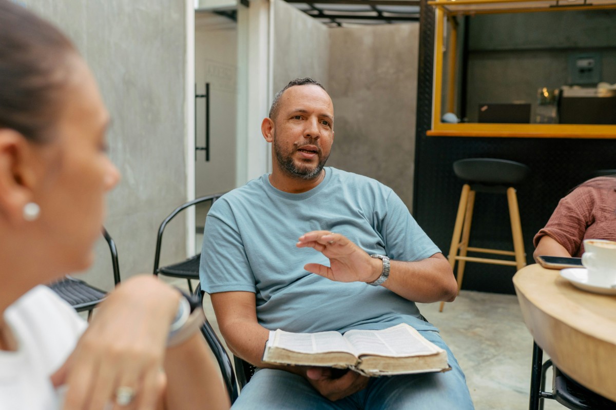 A man in a blue shirt holds an open book, gesturing while speaking to a woman in the foreground at a round table indoors.