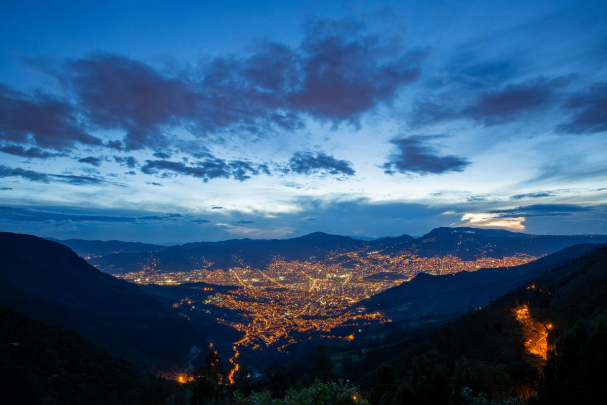 A cityscape at dusk, with glowing city lights and distant mountains under a partly cloudy sky.