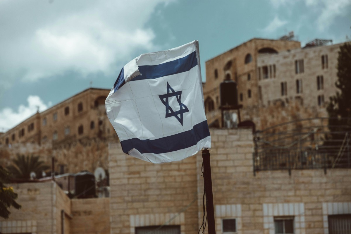 Israeli flag waving in front of stone buildings under a partly cloudy sky.