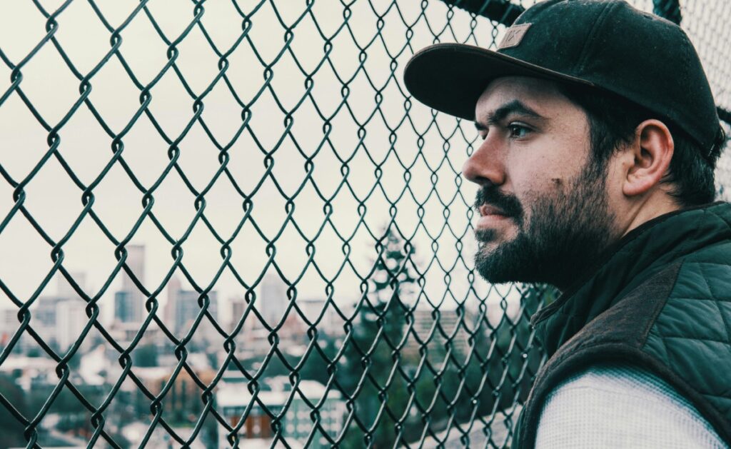 Man in a cap looking through a chain-link fence with a cityscape in the background.