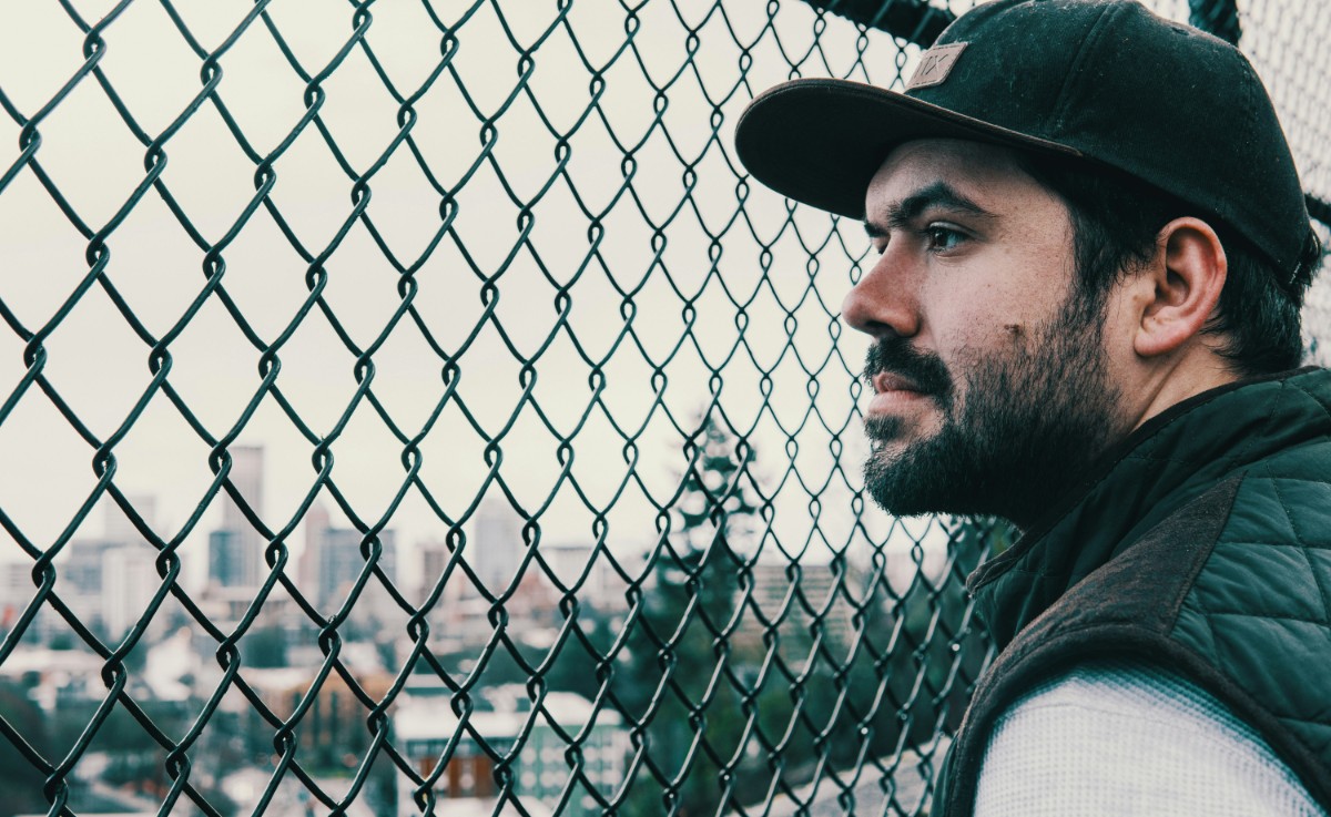 Man in a cap looking through a chain-link fence with a cityscape in the background.