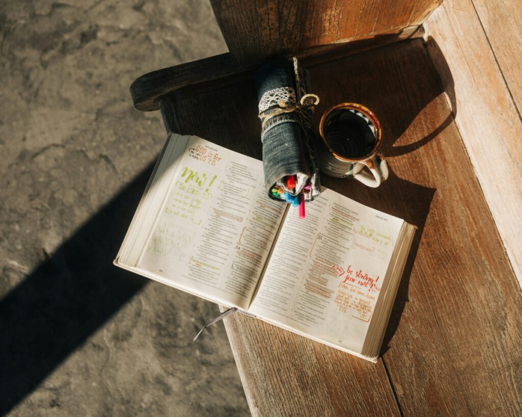 Open book with highlighted text on a wooden bench, accompanied by a rolled-up denim pouch and a cup of coffee.