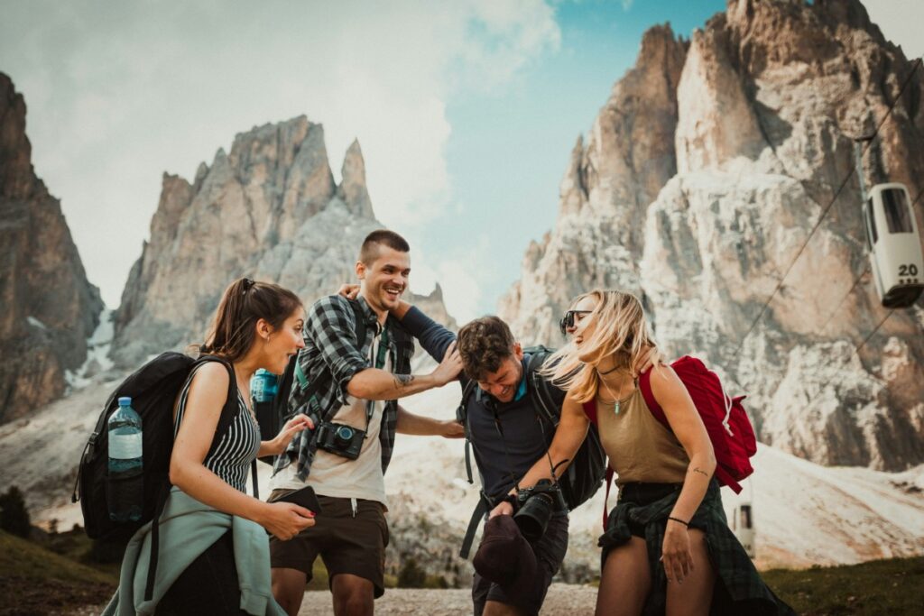 A group of five people with backpacks laugh and walk together in a mountainous landscape with rocky peaks in the background.