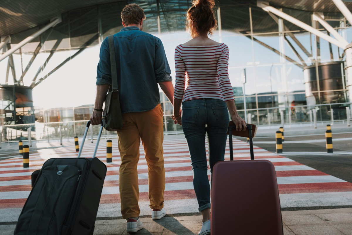 A couple holding hands, walking with suitcases towards a modern building with large glass windows and a red-and-white striped crossing.