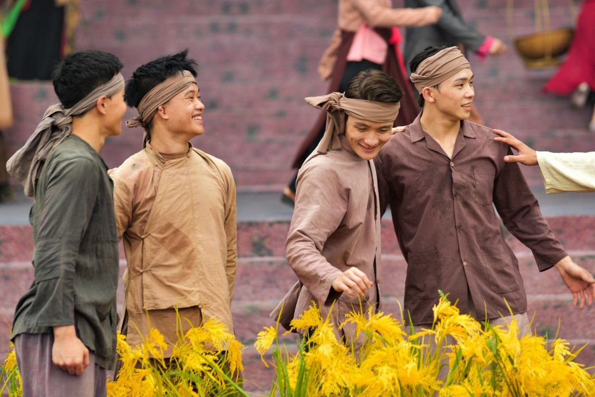 Four people wearing traditional attire and headbands stand next to yellow flowers, with one person pointing. Steps and blurred figures are in the background.