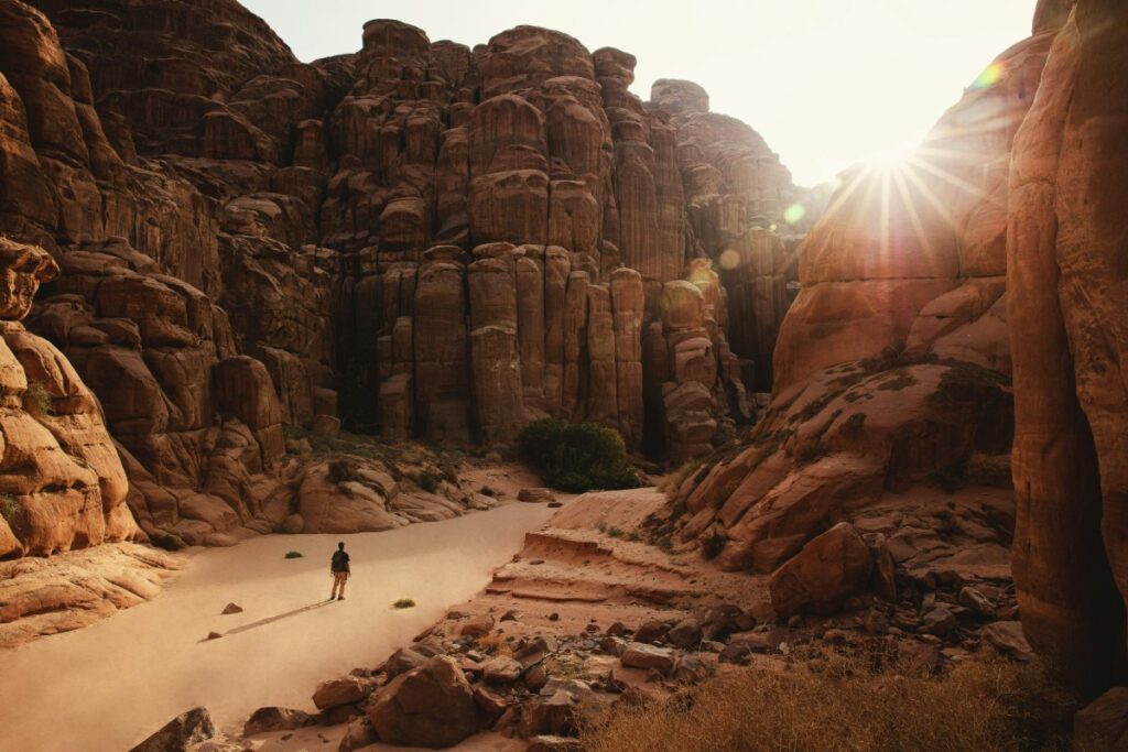 Person walking in a sunlit canyon with tall, rugged rock formations and a bright sunflare overhead.