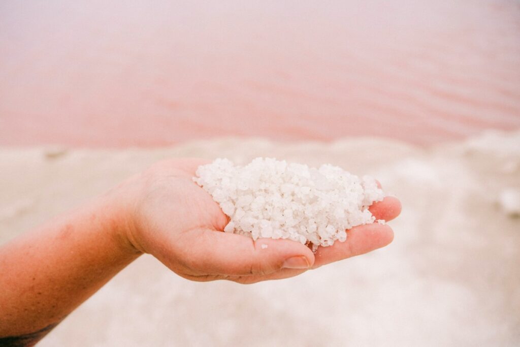 Close-up of a hand holding a pile of coarse white salt, with a pinkish body of water in the background.