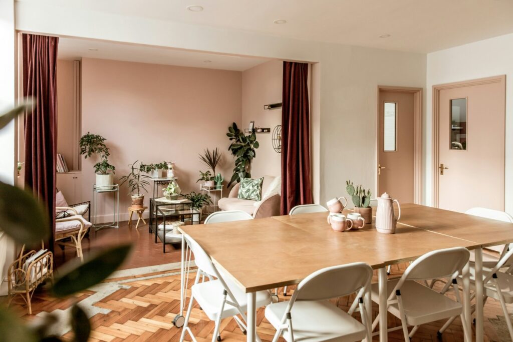 A dining area with a wooden table and white chairs, next to a living space with pink walls, plants, and chairs. There are burgundy curtains partially open between the spaces.