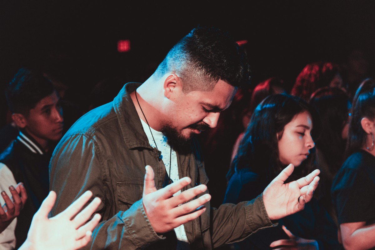 People standing with eyes closed and hands raised in a dark room, likely participating in a group activity or event.