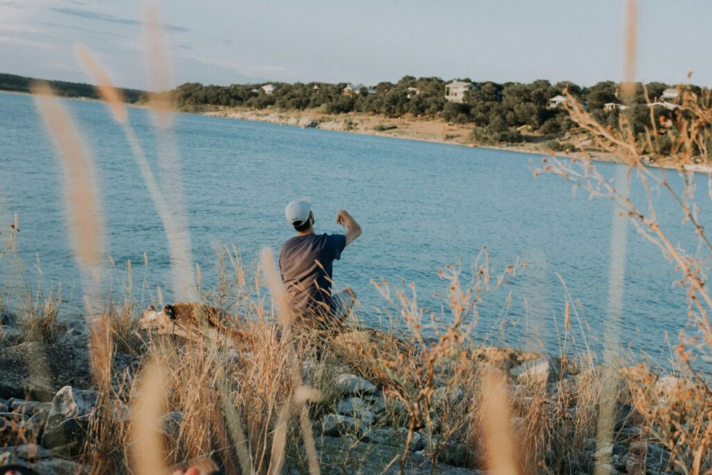 Person sitting on a rocky shore, waving toward a lake. Grassy plants in the foreground and a tree-lined horizon in the background.