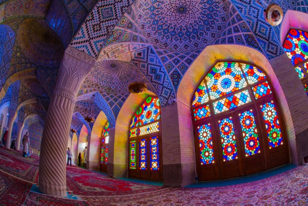 Interior of a mosque with intricate stained glass windows casting colorful light patterns on the walls and carpet, featuring ornate architectural designs on the ceiling and columns.