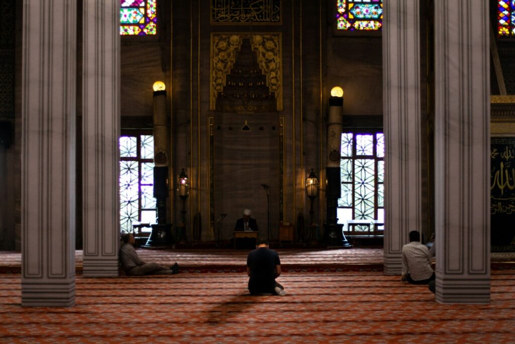 Interior of a dimly lit mosque with ornate columns and stained glass windows. People are seated on the red carpeted floor near the prayer area.
