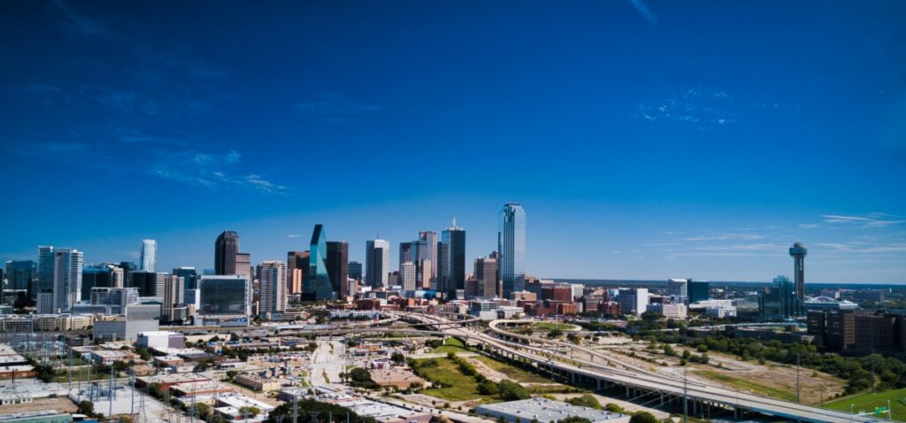 Aerial view of a city skyline with modern skyscrapers, clear blue sky, and surrounding roads and greenery.