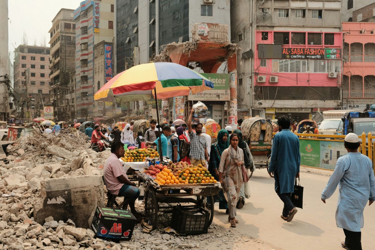 Street market with fruit vendors under umbrellas, surrounded by rubble and busy pedestrians. Buildings in the background.