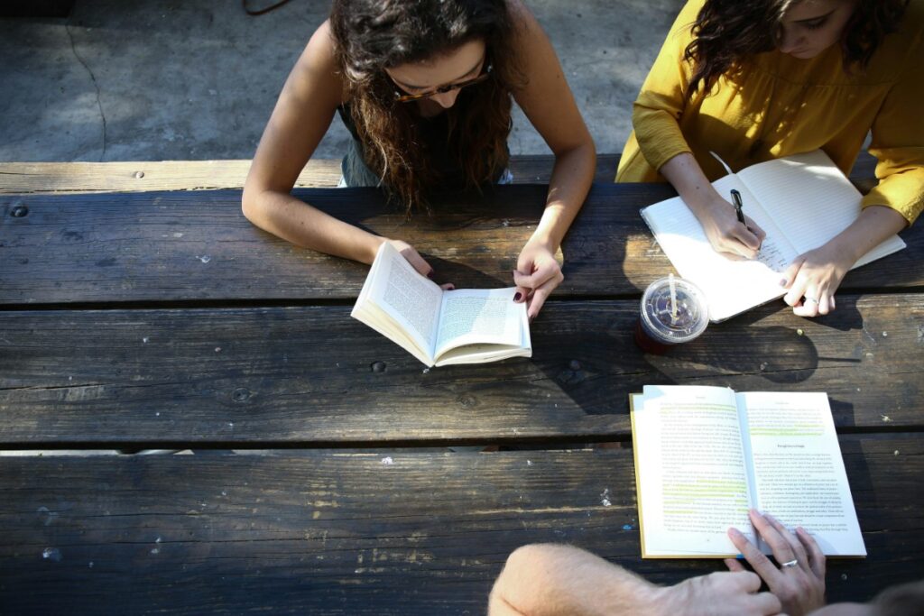 Three people sit around a wooden table; two read open books while one writes in a notebook. A plastic cup with a straw is on the table.