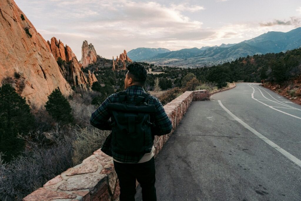 Person standing by a stone wall overlooking a road with rocky cliffs and mountains in the background.