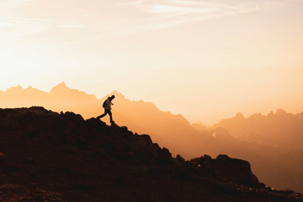 Person hiking uphill on a rocky terrain during sunset with mountains in the background.