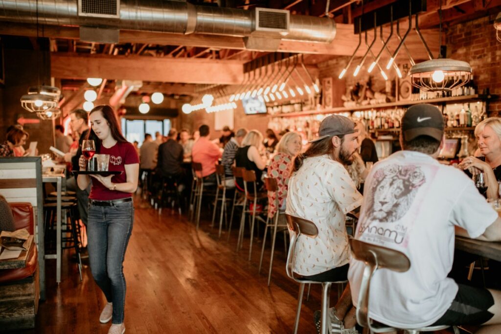 A busy bar scene with people sitting at a long counter. A server carries drinks, and ambient lighting creates a warm atmosphere.