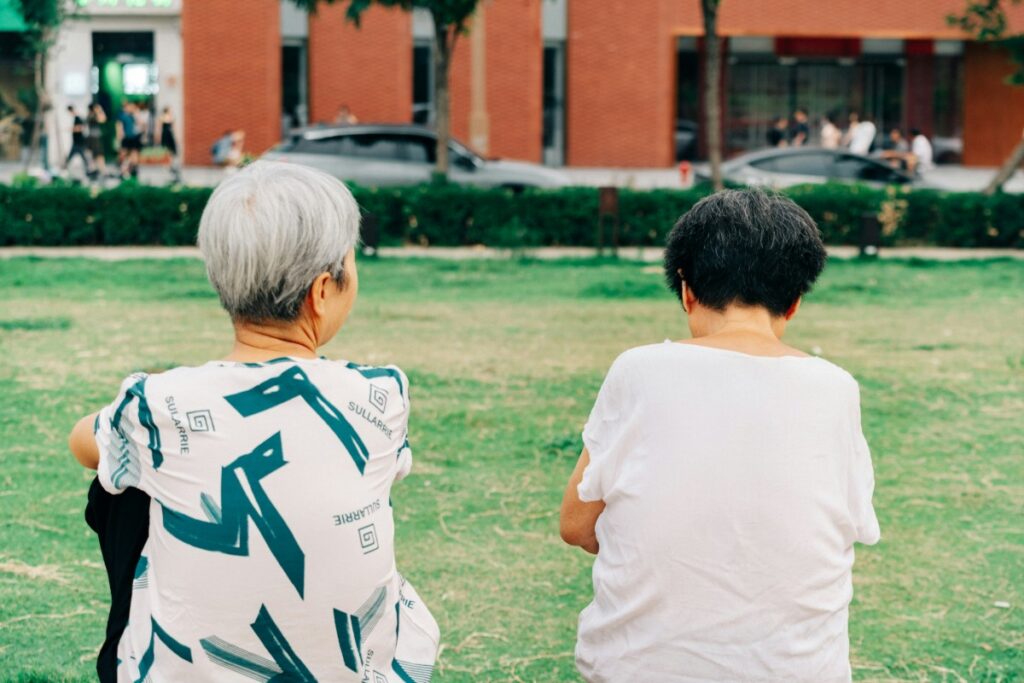Two people with short hair sit on a green lawn, facing away. A street and brick building are in the background.