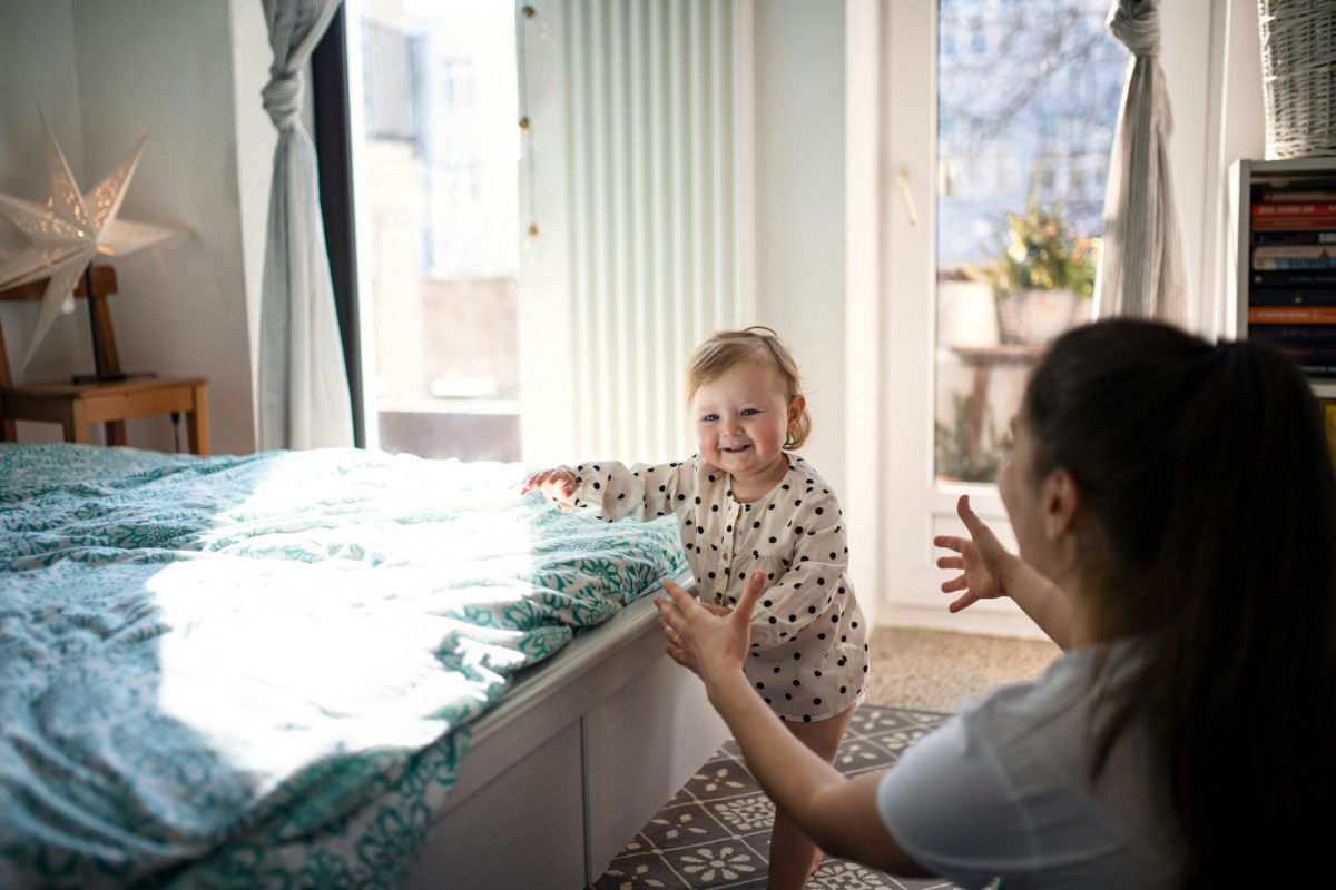 A baby in pajamas stands by a bed, smiling, while a woman kneels nearby with open arms. The room is bright with sunlight.