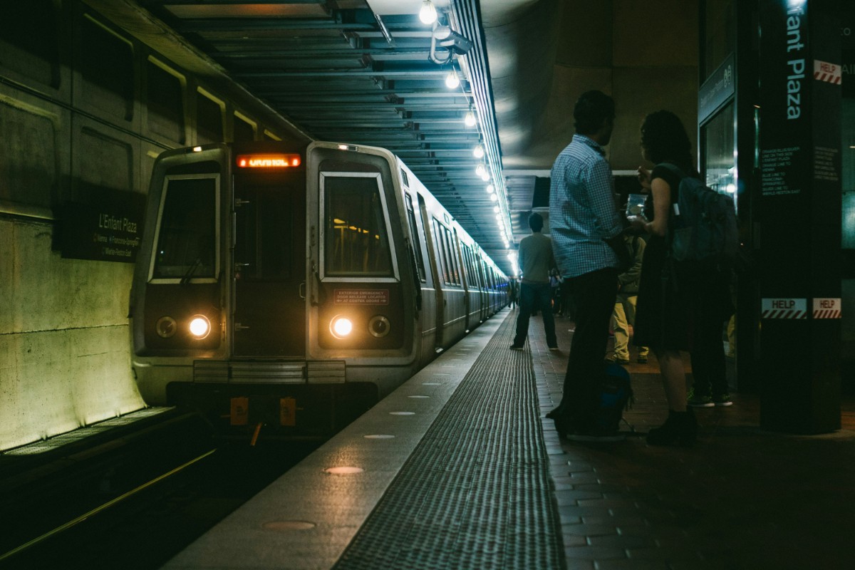 A subway train approaches the platform in a dimly lit underground station as two people stand nearby.