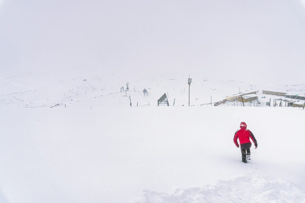 A person in a red jacket walks through a snowy landscape with a few structures and a ski lift in the background under an overcast sky.
