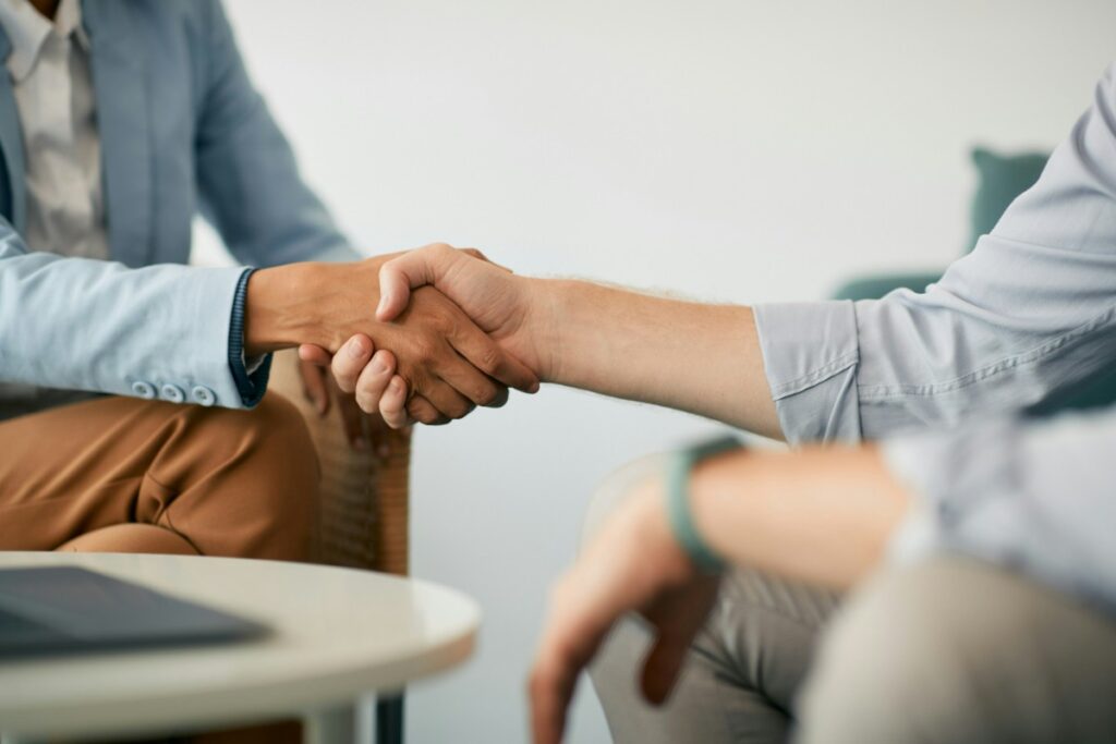 Two people shaking hands over a table with a laptop, one wearing a blue jacket, the other a light-colored shirt.