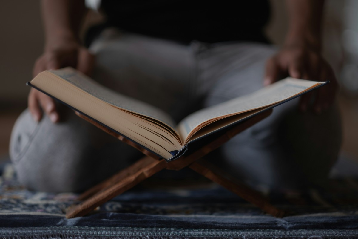 Person sitting cross-legged on a mat, holding an open book on a wooden stand.