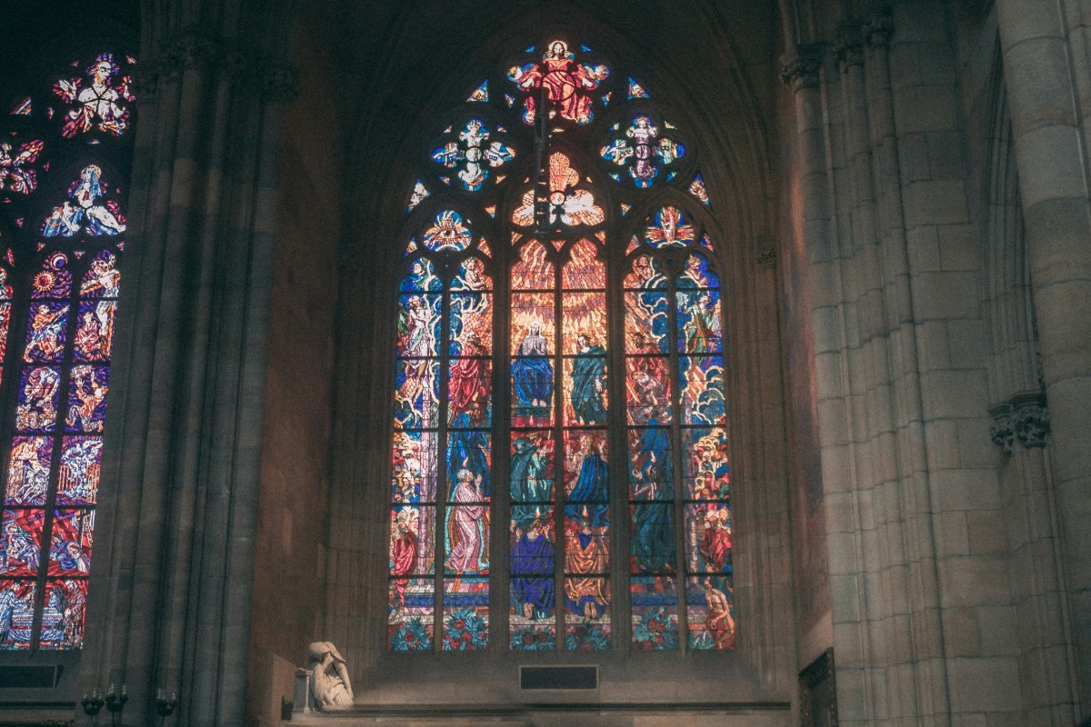 Stained glass windows in a church depict religious figures and scenes, with intricate designs and vibrant colors, surrounded by stone architecture.