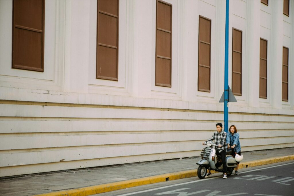 Two people ride a scooter along a street with a large white building and brown windows in the background.