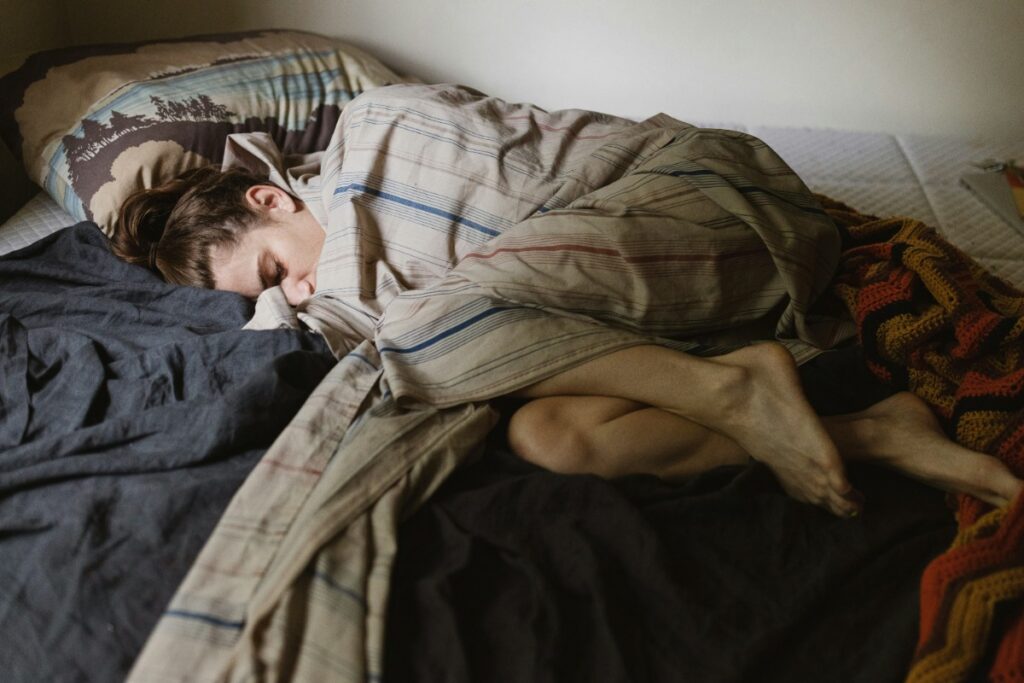 Person sleeping curled up on a bed, wrapped in a plaid blanket, with head resting on a pillow.
