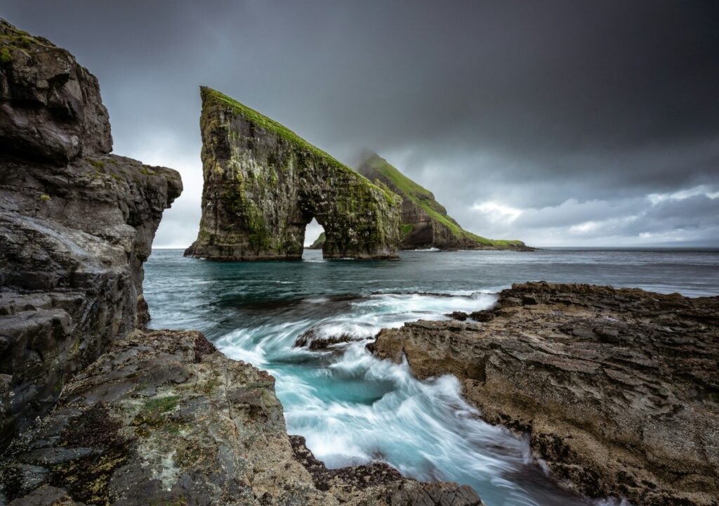 Dramatic seascape with towering cliffs and an arch over the sea, under a cloudy sky with rough waves crashing against rocks in the foreground.