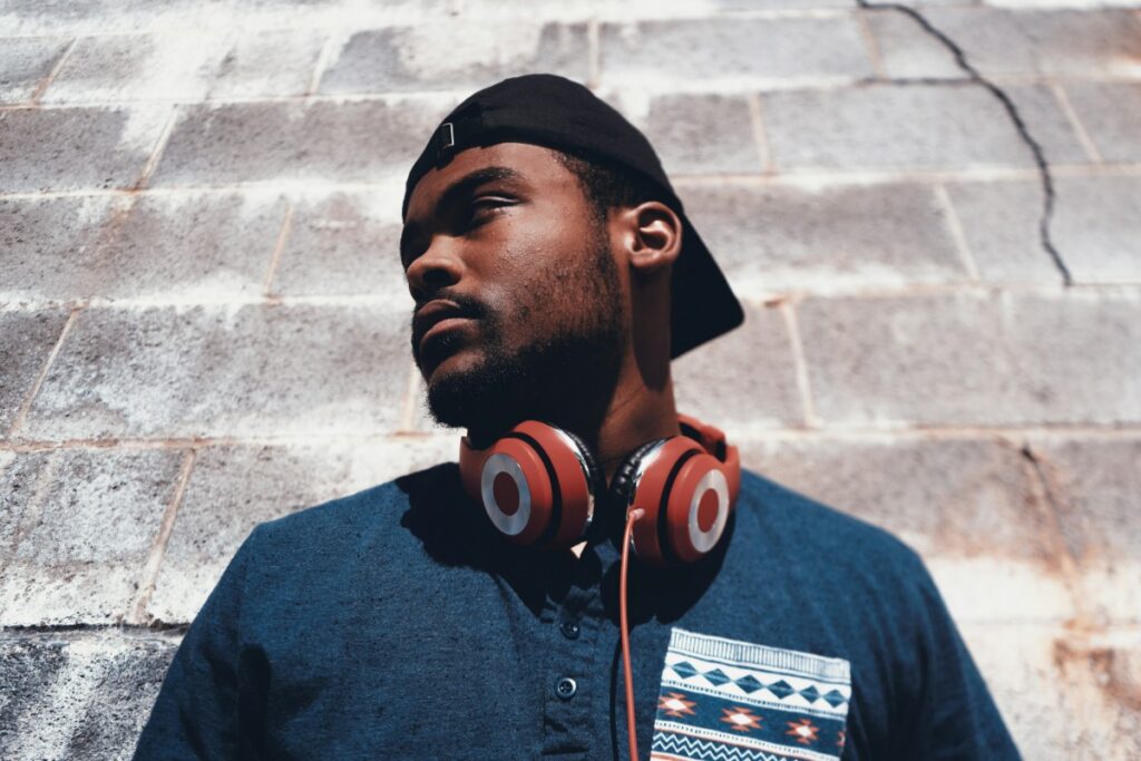 A person wearing a backward cap, blue shirt, and red headphones stands against a cracked brick wall, looking to the side.