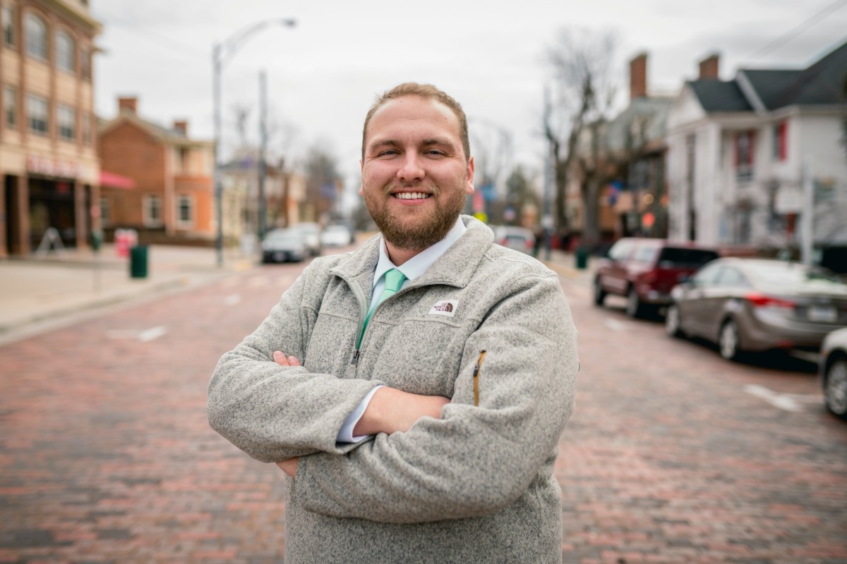A person stands smiling with arms crossed on a brick-paved street lined with parked cars and buildings in the background.