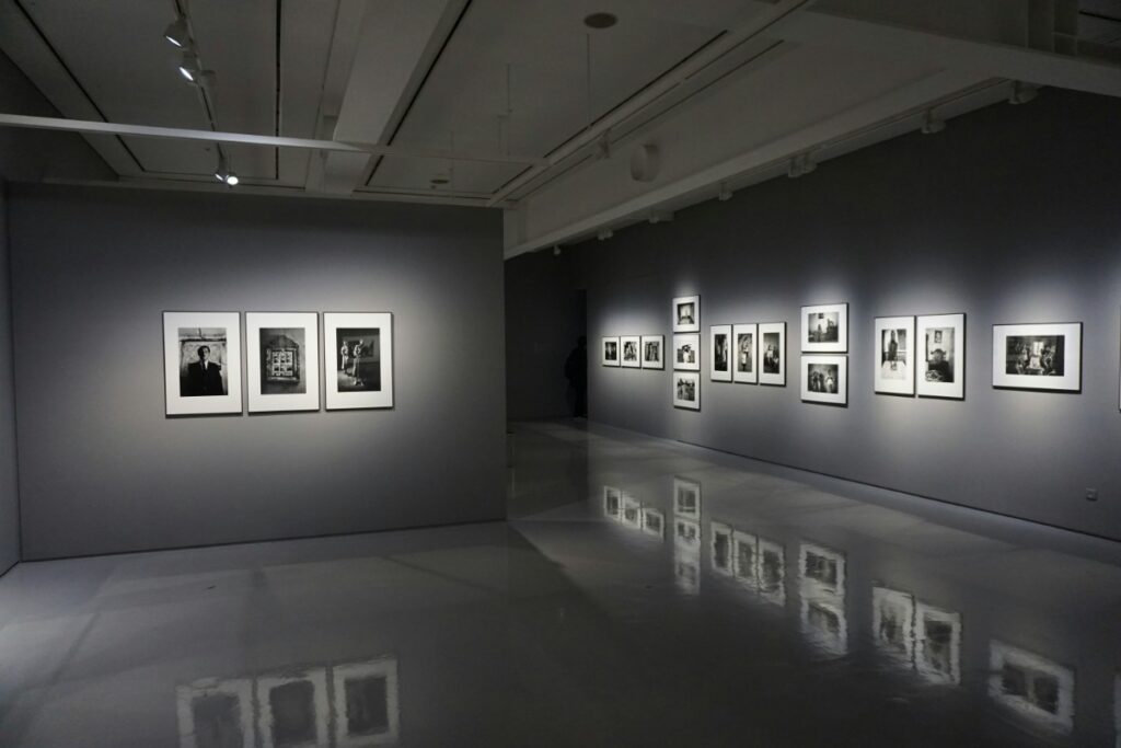 A dimly lit art gallery interior with framed black and white photographs displayed on grey walls, reflecting on a polished floor.