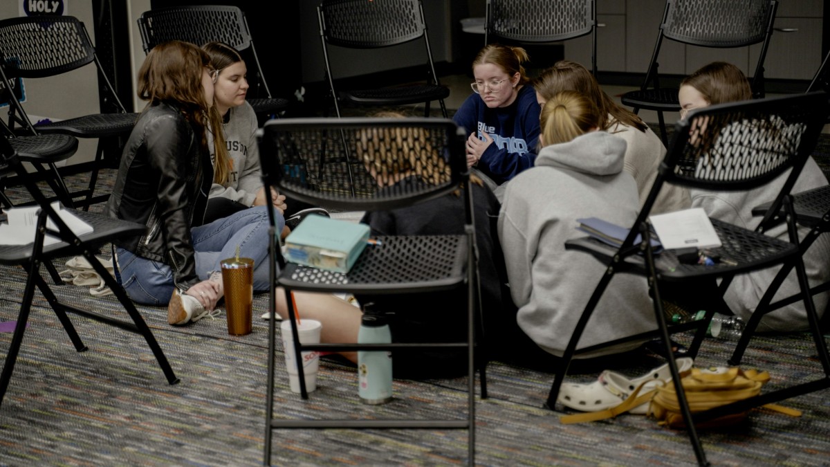 A group of people sit on the floor in a circle, surrounded by chairs, engaging in conversation.