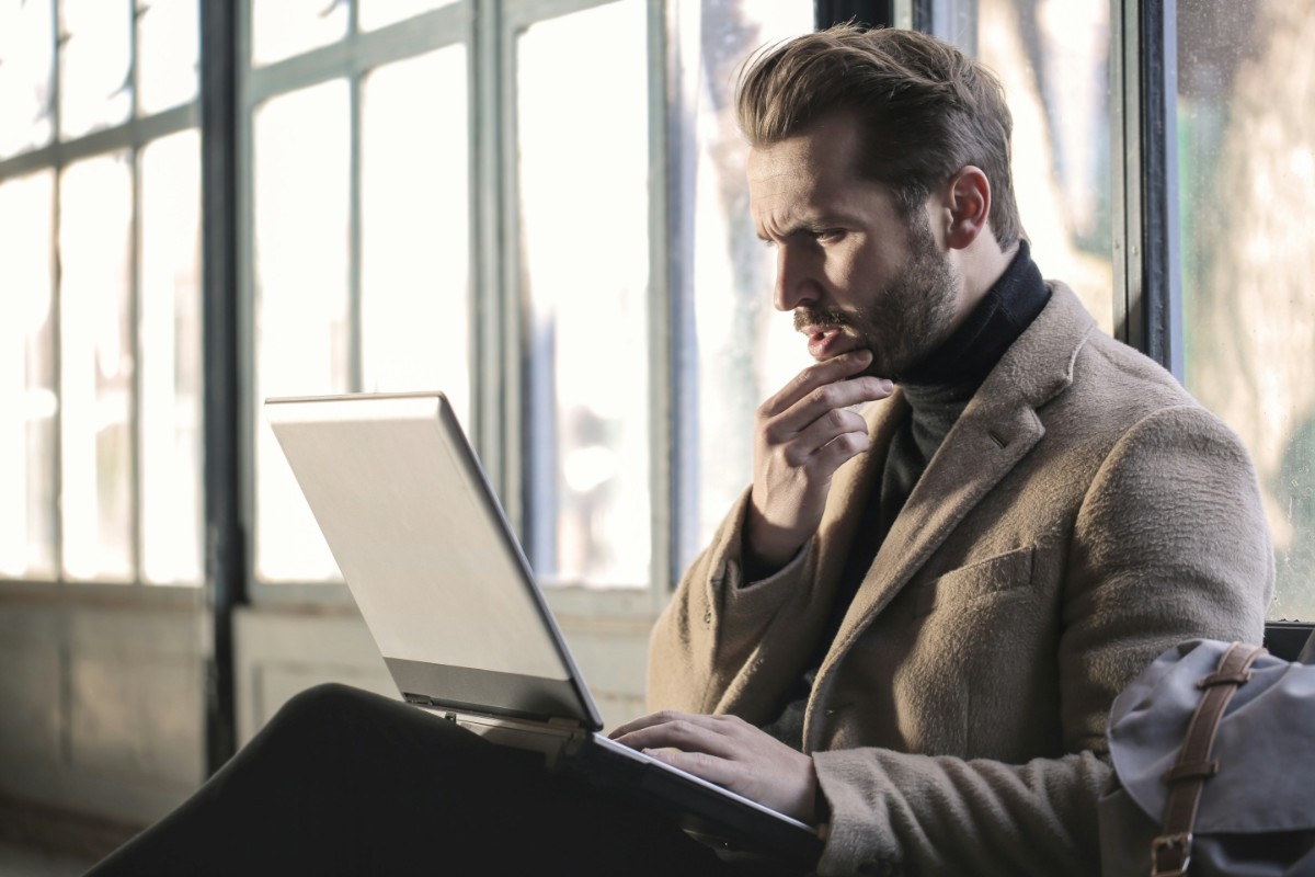 Man in a jacket sitting and looking thoughtfully at a laptop screen in a bright room.