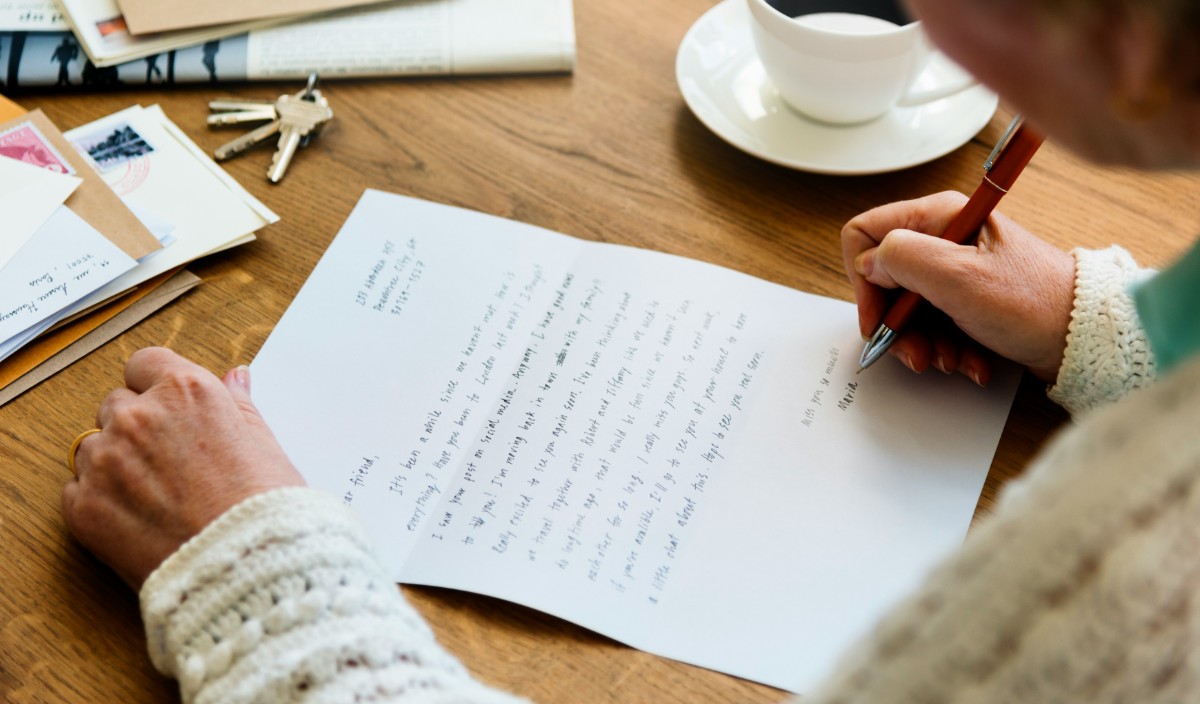 A person writing a handwritten letter at a wooden table, with envelopes, keys, and a cup nearby.