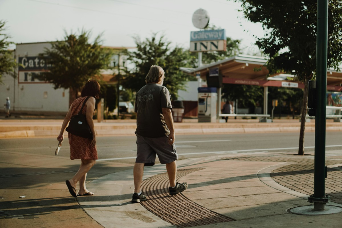 Two people crossing a street at a crosswalk in a small town area with buildings and trees in the background.