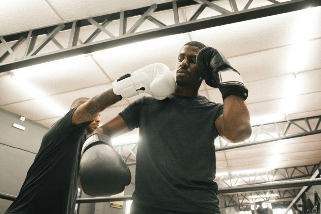 Two boxers sparring in a gym, one with white gloves making contact with the others head. The gym has mirrored walls and overhead lighting.