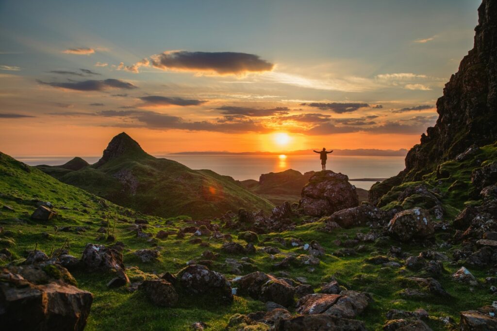 Person standing on rocky terrain at sunset, arms raised. Sky is orange and pink with scattered clouds. Green hills and distant water visible in the background.