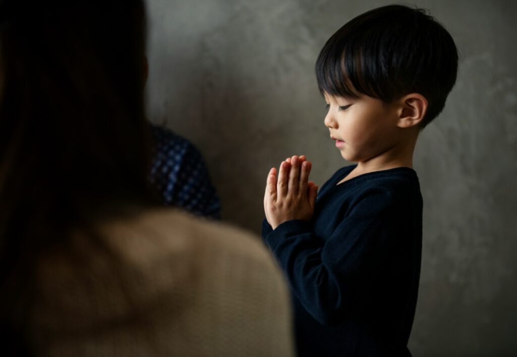 A young boy with dark hair, wearing a black shirt, stands with hands clasped in prayer, eyes closed, against a neutral background.