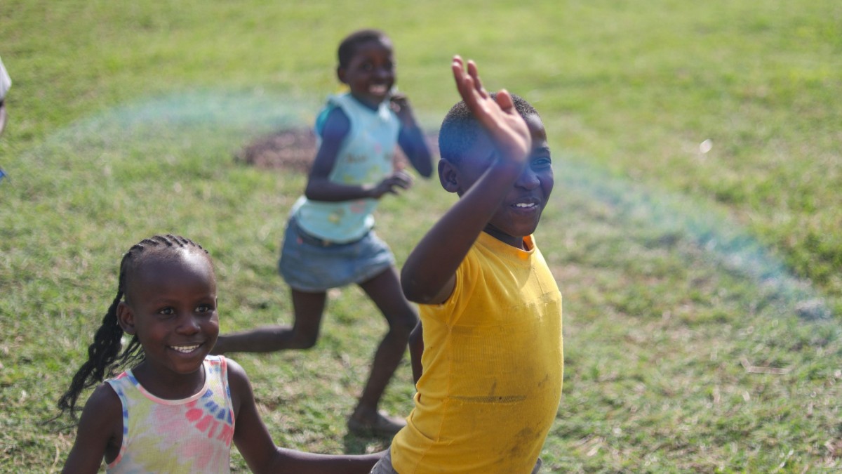 Children play and smile on a grassy field, with one child in a yellow shirt waving.