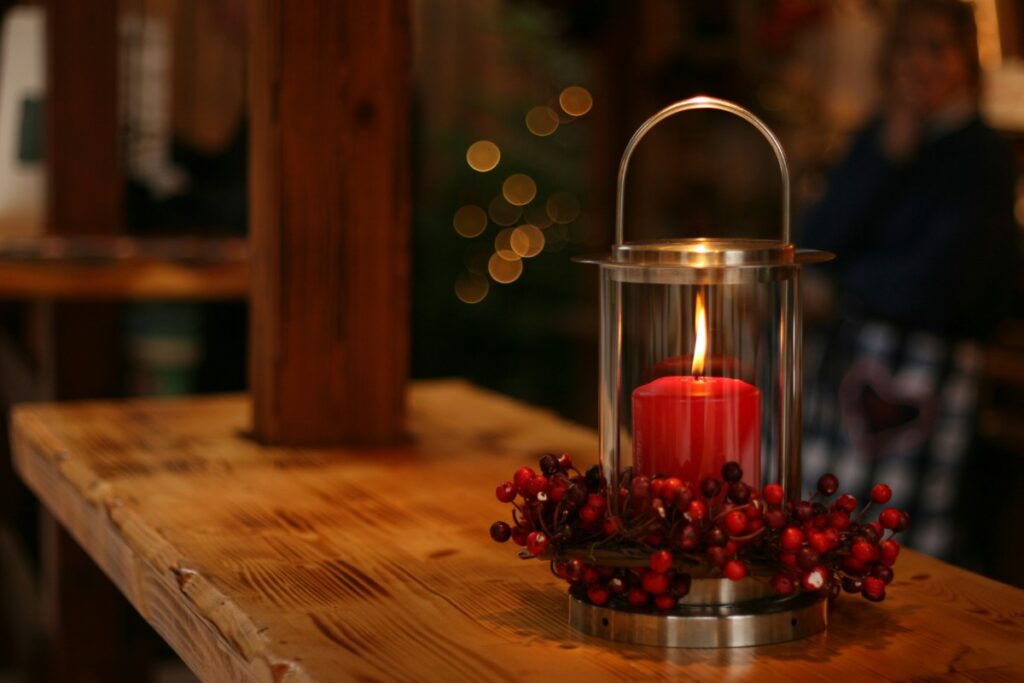 A red candle burns inside a glass lantern on a wooden table, surrounded by red berries. Blurred background with warm lighting.