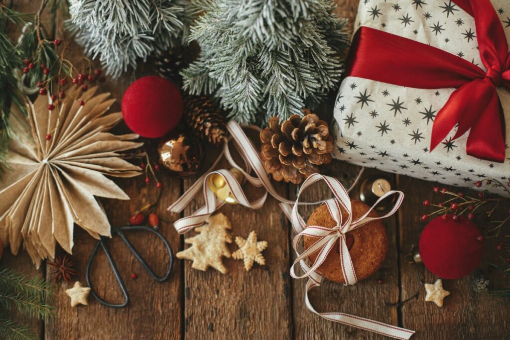 Christmas-themed arrangement with two small snowy trees, a wrapped gift, red ornaments, pinecones, stars, scissors, and decorative paper on a wooden surface.