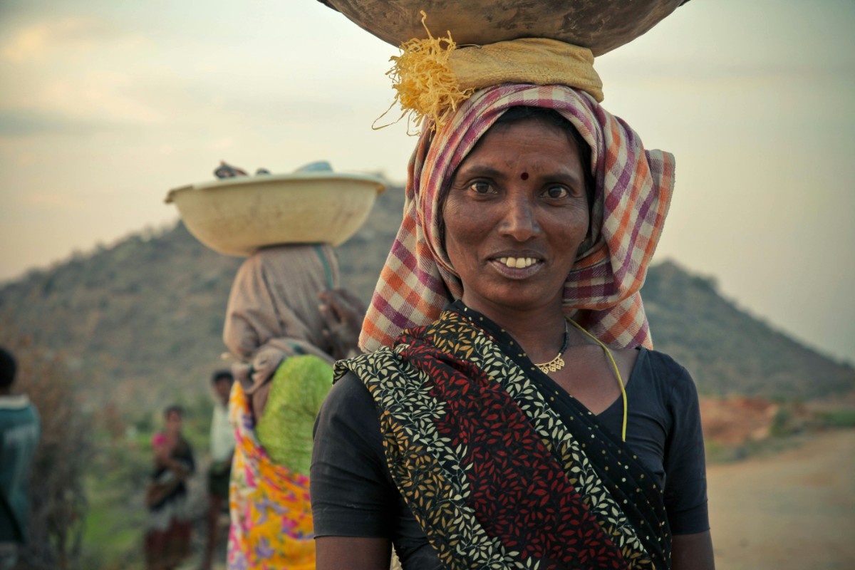 A woman smiling, carrying a basket on her head, stands outdoors with other people in the background.