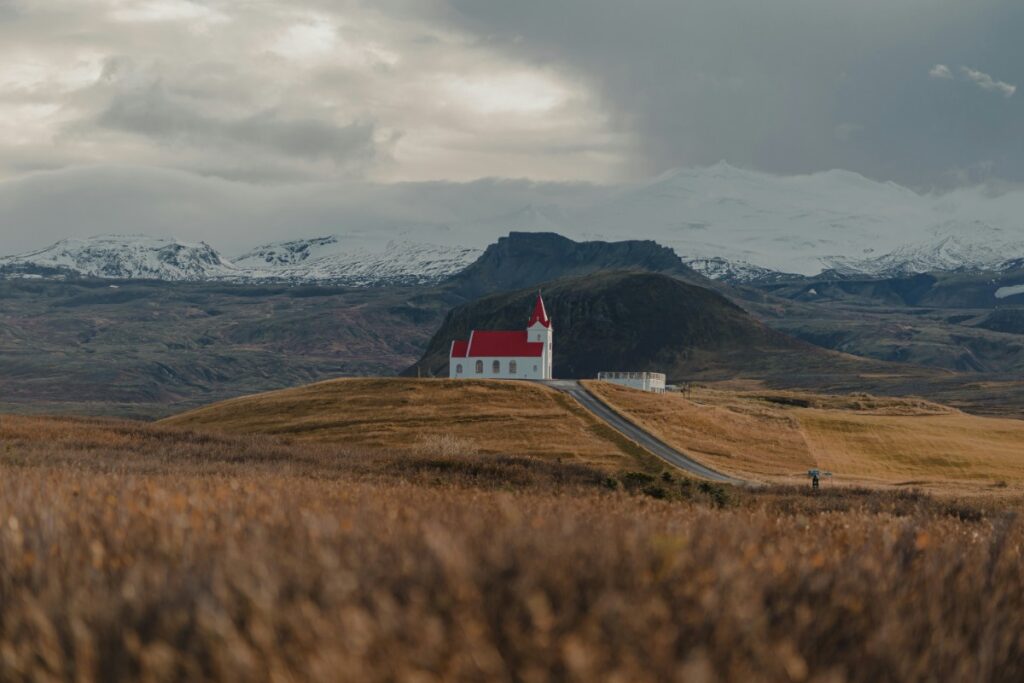A small white church with a red roof stands on a hill, surrounded by a vast landscape with mountains and cloudy skies in the background.