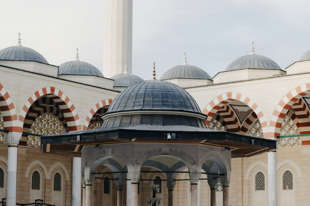 A mosque with multiple domes, arched facades, and a central courtyard. The architecture features red and white striped patterns.