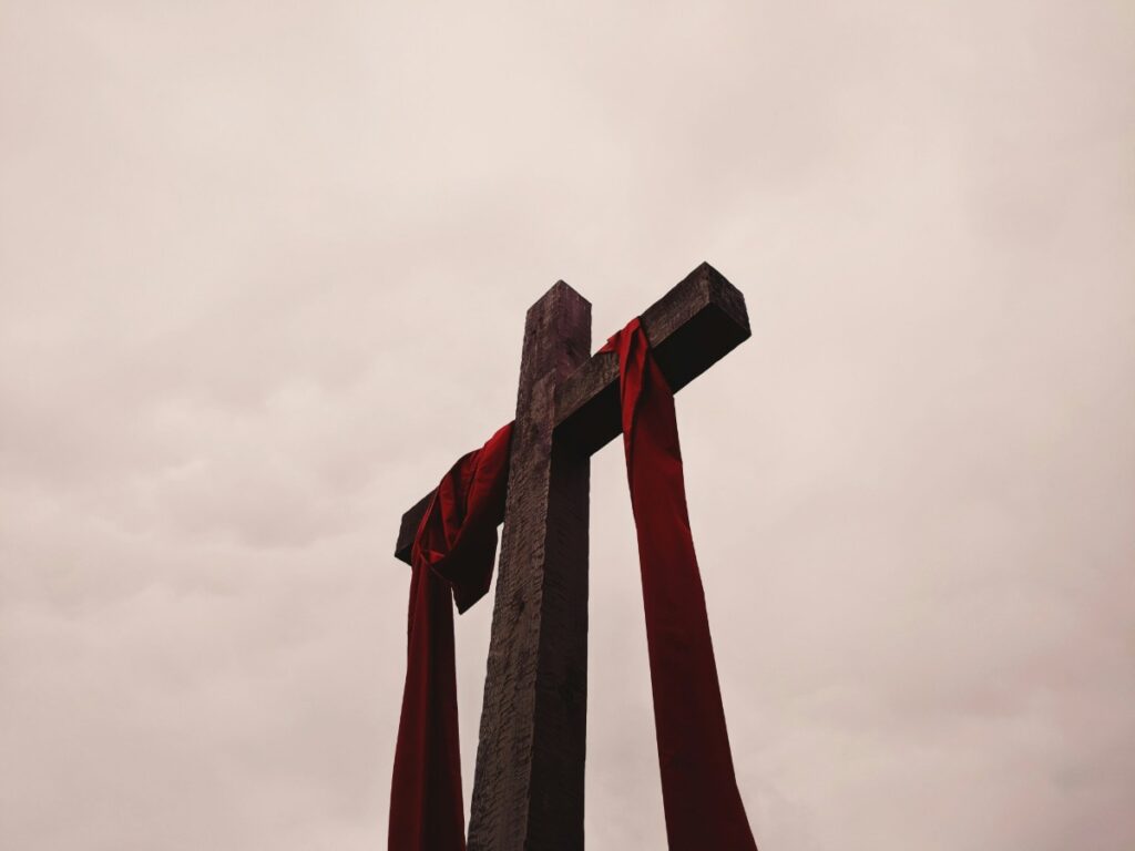 A wooden cross with red fabric draped over its arms against a cloudy sky.
