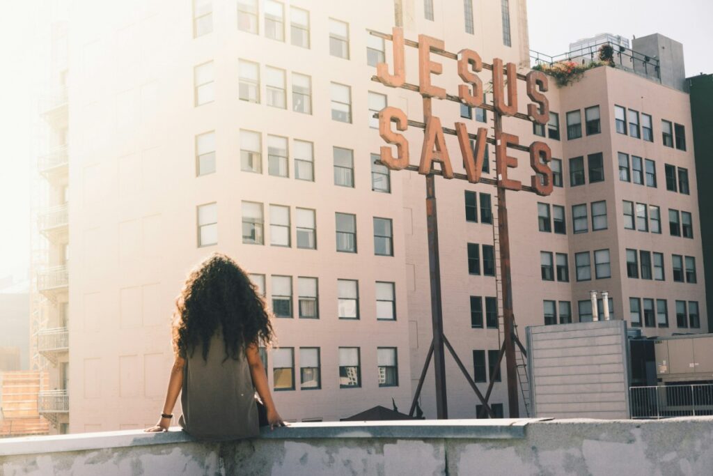 Person sitting on a rooftop ledge, facing a large JESUS SAVES sign on a tall building.